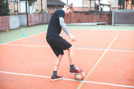 Young man focuses on controlling a football on an outdoor court. He carefully juggles the ball with precision, surrounded by an urban backdrop of buildings and a goal, combining sports and skill.の写真素材