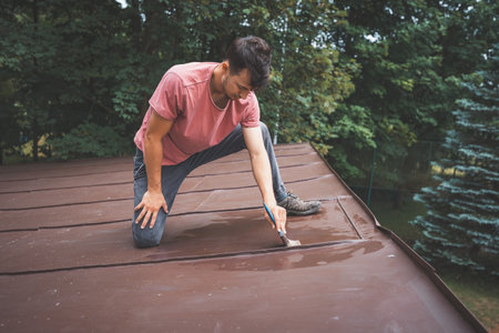 Man crouches down, carefully painting the surface of a roof with a small brush. His focus is on applying even strokes of paint. The outdoor setting is surrounded by trees, creating a peaceful atmosphere.の写真素材