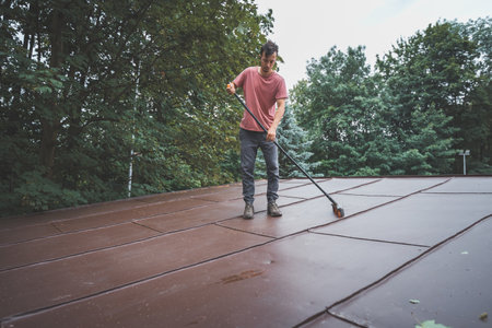 Man is cleaning a slanted roof with a long broom, surrounded by trees. The outdoor setting features lush greenery, with the individual focused on his task of removing debris from the roof.の写真素材
