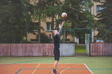 Basketball player prepares to shoot the ball on an outdoor court, positioning himself for a focused throw. The scene captures the athlete mid-motion, with a backdrop of trees and urban buildings.の写真素材