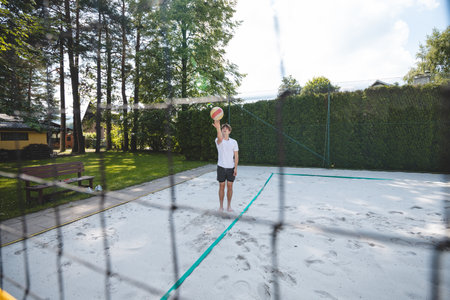 Young man tossing a volleyball into the air, preparing for a spike on a sand court. Captured from behind the net, showcasing a dynamic outdoor summer sports moment.の写真素材