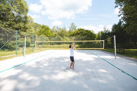 Young man tossing a volleyball into the air on a sandy court during a sunny day. The casual outdoor scene highlights fun, summer sports, and recreation in a natural setting.の写真素材