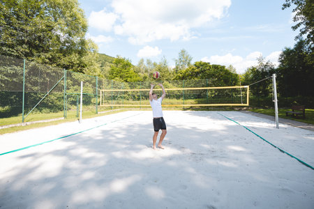 Young man tossing a volleyball into the air on a sandy court during a sunny day. The casual outdoor scene highlights fun, summer sports, and recreation in a natural setting.の写真素材