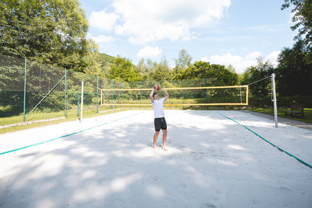 Young man tossing a volleyball into the air on a sandy court during a sunny day. The casual outdoor scene highlights fun, summer sports, and recreation in a natural setting.の写真素材