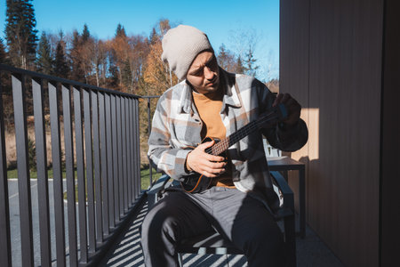 Young musician sits on a sunny balcony, immersed in playing the ukulele. Surrounded by autumn trees, he enjoys the peaceful moment outdoors, lost in his thoughts and musicの写真素材