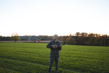 Musician in a plaid shirt and beanie plays the ukulele in a green field at sunset. The serene rural scene beautifully combines music, nature, and tranquility.の写真素材