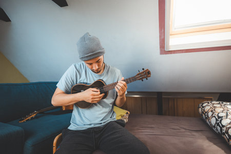 Musician with smile sits on a cozy bed, strumming a black ukulele. Enjoying the feeling of playing the ukulele and immersing oneself in the creative spirit.の写真素材