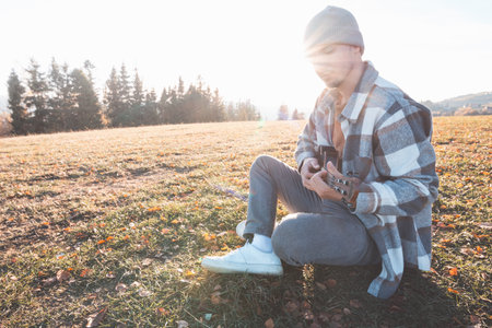Young man sits on a grassy field in the warm autumn sunlight, smiling as he plays the ukulele....