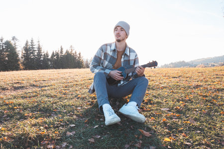 Young man sits on a grassy field in the warm autumn sunlight, smiling as he plays the ukulele. Surrounded by golden tones and distant trees, the moment reflects joy, creativity, and a deep connection.の写真素材