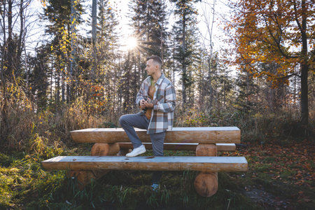 Musician stands by a rustic wooden bench in an autumn forest, playing the ukulele. With colorful leaves, a gazebo in the background, and a serene atmosphere, the scene reflects a peaceful connection.の写真素材
