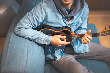 Musician in a denim jacket and hoodie sits comfortably on a blue sofa, playing a black ukulele. Boy with talent is playing the ukulele in the safe environment of his home. Focus on music.の写真素材
