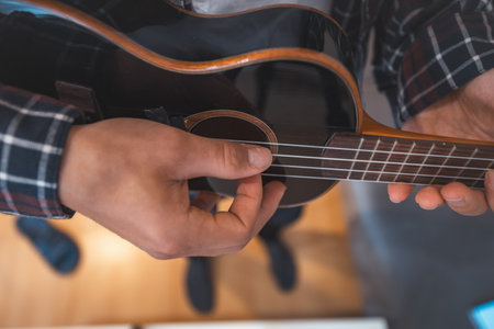 Close-up perspective captures a man's fingers plucking the strings of a glossy black ukulele. Making a music. Pleasure of playing an instrument.の写真素材