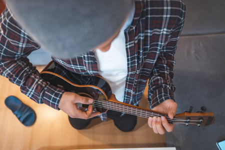 Close-up of a man's hands expertly gripping a ukulele, with one hand pressing the fretboard and the other poised to strum. His plaid shirt and the instrument's wooden frame add character.の写真素材