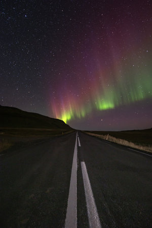 Mesmerizing aurora borealis over a quiet road on Snaefellsnes peninsula, West Iceland. Green and pink hues light up the starry night, creating a surreal landscape.の写真素材