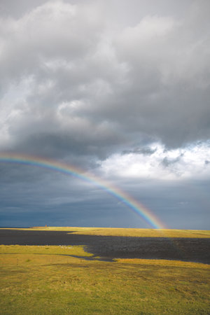 Vivid rainbow stretches across the cloudy sky above a serene Icelandic meadow. Snaefellsnes peninsula, west Iceland. Vibrant colors contrast beautifully with the soft, moody clouds and golden grass.の写真素材