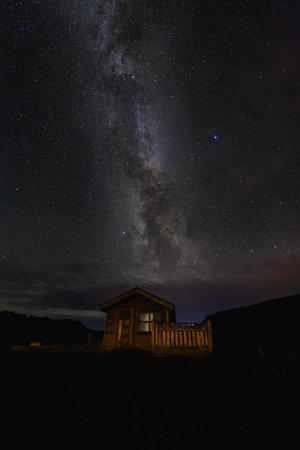 Cozy wooden cabin sits under a breathtaking night sky filled with the Milky Way galaxy's brilliance....
