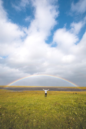 Person stands with arms wide open on a lush green meadow under a rainbow arch in Iceland. The...