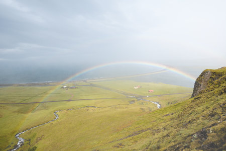 Vivid rainbow stretches across the cloudy sky above a serene Icelandic meadow. Snaefellsnes peninsula, west Iceland. Vibrant colors contrast beautifully with the soft, moody clouds and golden grass.の写真素材
