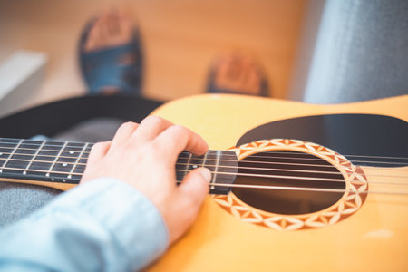 Relaxing on a cozy sofa, a man gently strums his acoustic guitar, lost in music. A peaceful home setting, soft lighting, and artistic inspiration create a warm, creative atmosphere.の写真素材