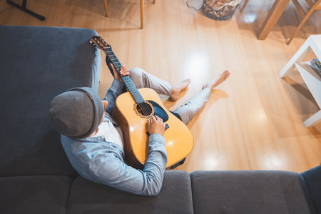 Musician sits on a gray couch, casually strumming an acoustic guitar, immersed in a moment of relaxation and introspection, creating a peaceful and creative atmosphere. Top view.の写真素材
