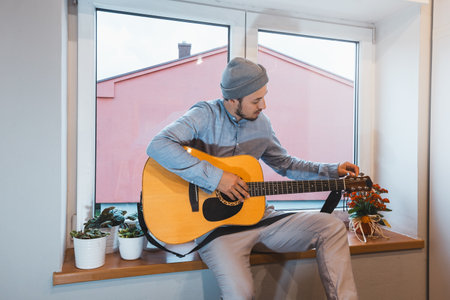 Musician sits on a windowsill, carefully tuning his acoustic guitar, surrounded by small plants and...