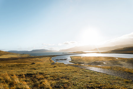 Golden light spills over the landscape behind Kirkjufellfoss towards Grundarfjordur, painting hills, rivers, and coastline in warm tones under a soft, glowing Icelandic sky.の写真素材