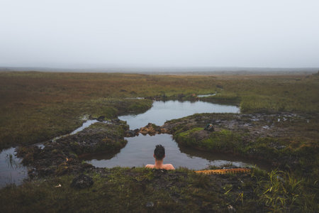 Young man lies in a natural Icelandic hot spring, surrounded by a misty landscape, in a moment of...