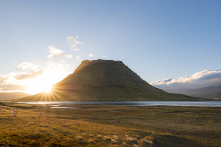 Kirkjufell mountain at sunrise with golden grasslands and soft clouds above, creating a peaceful and...