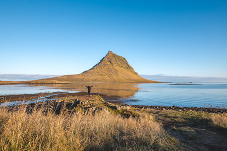 Lone traveler stands near the water gazing at Kirkjufell on Snaefellsnes peninsula Iceland as golden light and reflections shape a peaceful morning moment in nature.の写真素材