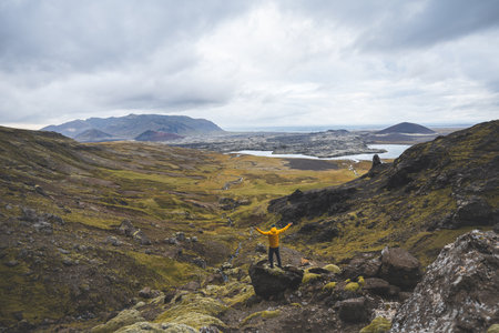 Hiker in yellow jacket stands above mossy rocks and volcanic valley on Snaefellsnes peninsula in Iceland, looking over lava fields and winding river beneath cloudy sky.の写真素材