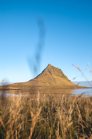 Iconic peak of Kirkjufell rises sharply on Snaefellsnes peninsula Iceland bathed in golden sunlight with a still lake and cloudless sky completing this perfect view.の写真素材