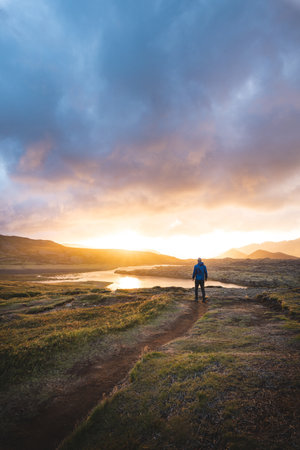 Lone traveler walks through the mossy lava fields of Berserkjahraun towards Selvallavatn lake, bathed in golden sunset light on Icelands Snaefellsnes Peninsula.の写真素材