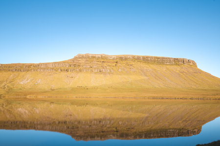 Perfectly still lake reflects the golden slopes of a plateau in western Iceland under a clear blue...