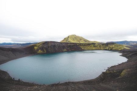 Serene volcanic lake with a tiny mossy island sits in the Icelandic highlands, surrounded by rugged hills and untouched nature under a soft overcast sky. Highlands in central Iceland.の写真素材