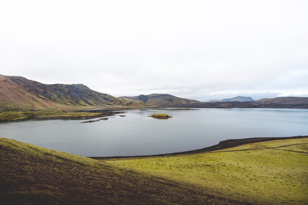 Serene volcanic lake with a tiny mossy island sits in the Icelandic highlands, surrounded by rugged hills and untouched nature under a soft overcast sky. Highlands in central Iceland.の写真素材