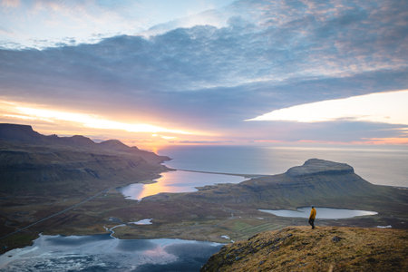 Hiker in a yellow jacket embraces the rising sun atop Kirkjufell Mountain, Iceland. Capturing a...