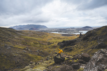 Hiker in yellow jacket stands above mossy rocks and volcanic valley on Snaefellsnes peninsula in Iceland, looking over lava fields and winding river beneath cloudy sky.の写真素材