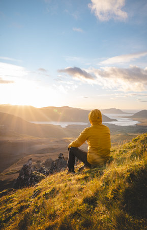 Traveler in a yellow jacket sits on the summit of The Horn on Snaefellsnes peninsula in Iceland watching the sun set over volcanic valleys and peaceful lakes below.の写真素材