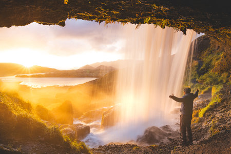 Man enjoys the view of a magical sunset from behind the Selvallafoss waterfall on the Snaefellsnes Peninsula in western Iceland, standing beneath the cold, clear water. Nature treasure.の写真素材