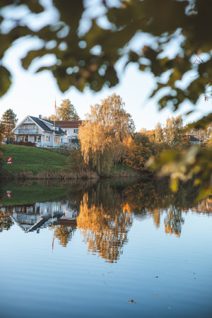 Autumn reflections dance on the still waters of Holmendammen Lake in Oslo, surrounded by colorful trees and hillside homes under a crisp, cloudless sky.の写真素材