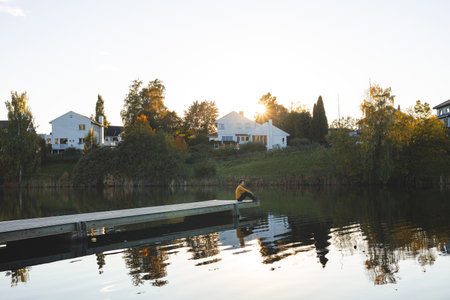 Lone man walks on pier an Oslo lake, Holmendammen as the sun sets behind charming houses, capturing a peaceful moment of reflection and stillness in Norway autumn magic.の写真素材