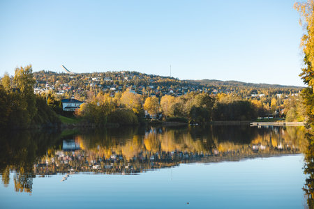 Autumn reflections dance on the still waters of Holmendammen Lake in Oslo, surrounded by colorful trees and hillside homes under a crisp, cloudless sky.の写真素材