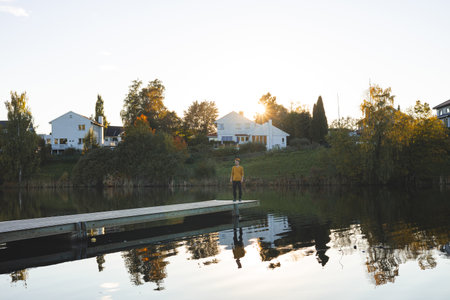 Lone man walks on pier an Oslo lake, Holmendammen as the sun sets behind charming houses, capturing a peaceful moment of reflection and stillness in Norway autumn magic.の写真素材