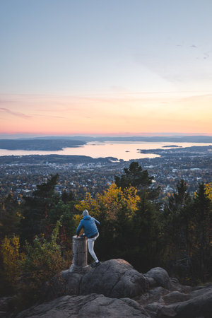Young traveler stands on a stump overlooking Oslo slowly falling asleep under the setting sun, as everything turns orange and pink. View from Vettakolltoppen, Norwegian nature.の写真素材