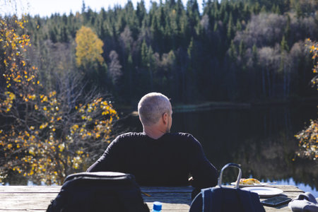 50 years old man relaxes by a forest lake in Oslo Marka, enjoying lunch under golden trees and sunlight, surrounded by still water and deep Scandinavian calm.の写真素材