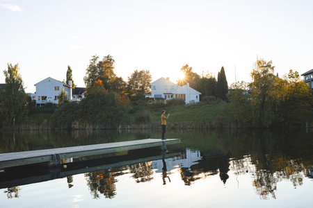 Lone man walks on pier an Oslo lake, Holmendammen as the sun sets behind charming houses, capturing a peaceful moment of reflection and stillness in Norway autumn magic.の写真素材