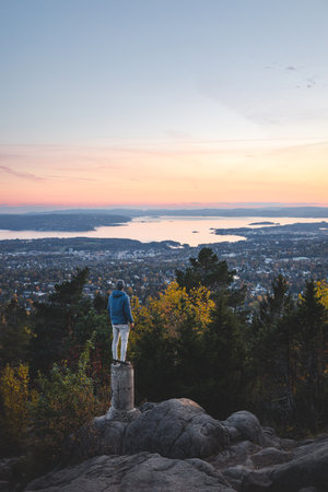 Young traveler stands on a stump overlooking Oslo slowly falling asleep under the setting sun, as everything turns orange and pink. View from Vettakolltoppen, Norwegian nature.の写真素材