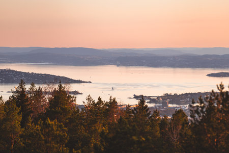 Soft pastel sunset over Oslofjord with islands, calm water and forested hills, reflecting the gentle Scandinavian evening mood and golden serenity.
