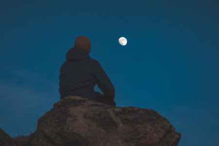 Lone traveler sits on rocks beneath the moon in Norway clear night sky. A poetic moment of solitude, thought, and northern minimalism in natureの写真素材
