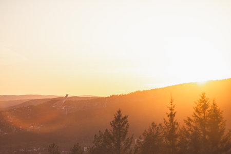 Holmenkollen ski jump rises through golden haze above Oslo pine-covered hills during sunset, glowing in warm Scandinavian spring atmosphere.の写真素材
